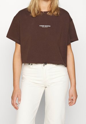 Osoba nosząca brązową krótką koszulkę z napisem "WORST BEHAVIOR" i białe spodnie z wysokim stanem, stojąca na jednolitym tle.