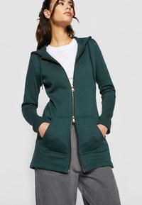 Sudadera verde con cremallera, capucha, cremallera dorada, dos bolsillos frontales y una textura suave. Combinada con pantalones grises de diseño casual.