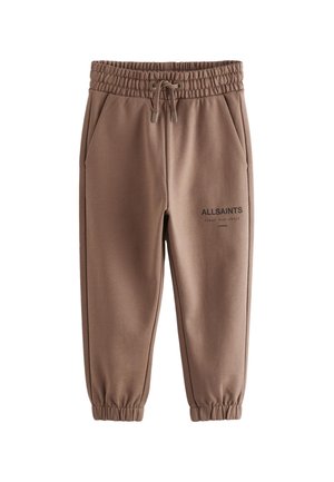 Dreng med fletninger iført beige AllSaints T-shirt, brune joggere og hvide sneakers, står med den ene hånd i lommen.