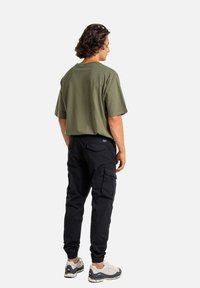 Mann mit lockigem Haar steht mit dem Rücken zur Kamera, trägt ein olivgrünes Oversized-T-Shirt, schwarze Cargo-Hosen und weiße Sneakers vor einem weißen Hintergrund.