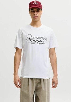 Mężczyzna w białej koszulce z czarnym nadrukiem w formie pisma oraz czerwonej czapce, stojący na tle jednolitego jasnego tła.