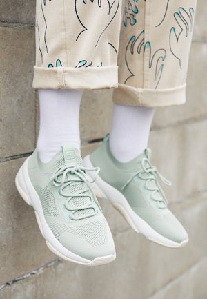Beine in cremefarbenen Hosen mit schwarzen und türkisfarbenen Handmustern, hochgekrempelt über weißen Socken und hellgrünen gestrickten Sneakern vor einer Steinmauer.