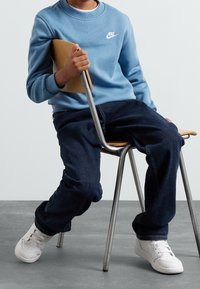 Barn i blå Nike-tröja, mörka jeans och vita sneakers sitter på sniskan på en trästol med metallben på en grå golv.