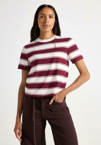 Burgunder- og hvide stribede T-shirt med rund hals og korte ærmer. Blødt bomuldsblandingsstof. Modellen har mørkebrun bukser på og står med hænderne i lommerne.