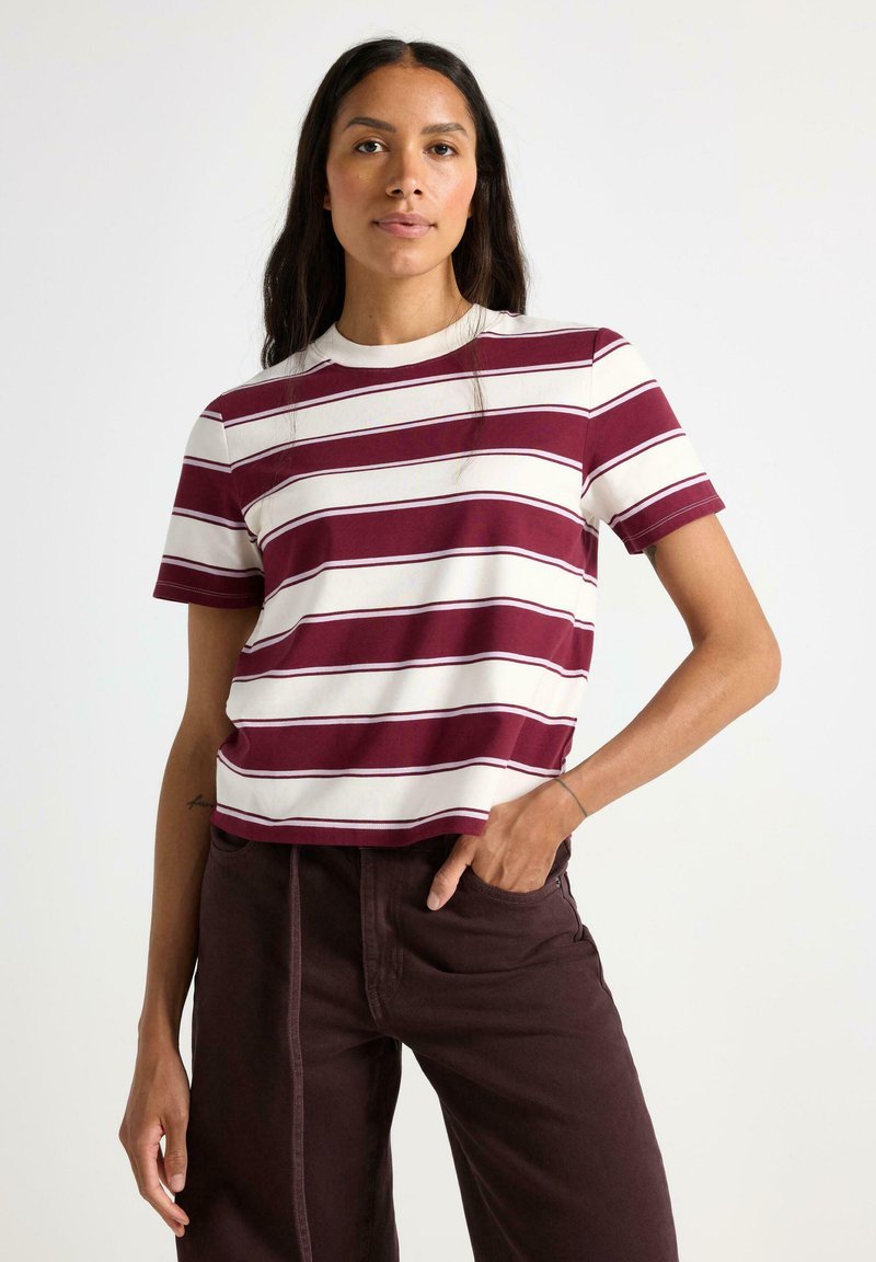 Burgunder- og hvide stribede T-shirt med rund hals og korte ærmer. Blødt bomuldsblandingsstof. Modellen har mørkebrun bukser på og står med hænderne i lommerne.