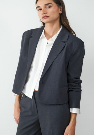 Donkergrijze blazer met een enkele knoop, notch revers en voorzak, over een wit shirt. Gladde textuur met een getailleerde pasvorm.