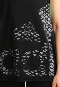 Černý bezrukávový top s texturovaným světle šedým grafickým designem s abstraktními tvary a nápisy na přední straně.