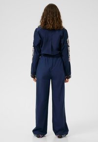 Salopetă bleu închis, cu o croială lejeră, decorată cu broderie florală pe mâneci și pe mansete, și o talie elastică pentru confort.