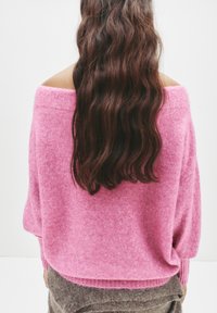 Roze oversized sweater met een off-the-shoulder ontwerp, geribbelde manchetten en een zachte textuur, gecombineerd met een bruine rok.