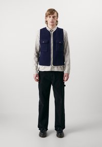 Jūras spēku vilnas bez piedurknēm veste ar divām priekšējām kabatām, ar rāvējslēdzēju un krēmkrāsas apdari, uzvilkta virs raibās krekliem un saskaņota ar melniem biksēm un kurpēm.