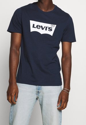 Uomo con una t-shirt Levi's blu navy con logo bianco e jeans azzurri, in piedi davanti a uno sfondo bianco uniforme.