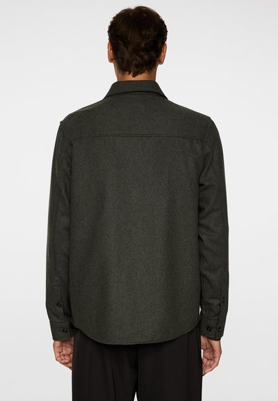 Mörkgrön ullskjorta med långa ärmar, krage och knäppning vid cuffs. Har en något rundad nederkant och en slät yta.