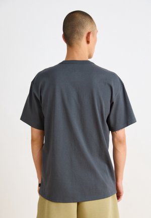 Homme à la tête rasée portant un t-shirt uni gris foncé à manches courtes et un short beige, de dos sur fond blanc.