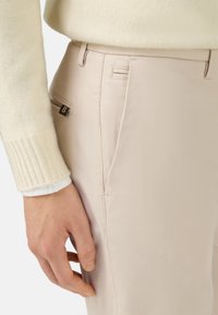 Pantalones beige claros hechos de un tejido suave, con un bolsillo lateral y costuras mínimas. Mano apoyada en la cintura.