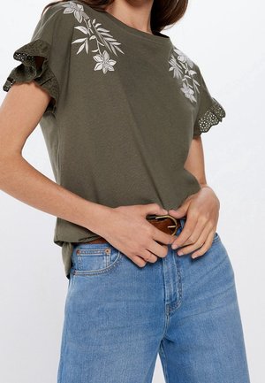 Mujer con blusa verde oliva con bordado floral blanco y mangas con volantes de encaje, metida dentro de unos vaqueros azul claro con un cinturón marrón.