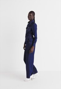Salopetă din denim de culoare albastru închis cu un top ajustat, pantaloni cu croială largă, guler ascuțit și buzunare frontale. Modelul poartă tocuri albe.