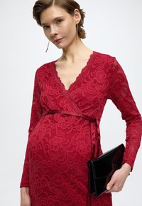 Piros csipkés, hosszú ujjú, V-nyakú, szatén övvel ellátott átlapolós ruha; virágmintás és egy fényes fekete borítéktáska látható, amit a deréknál tartanak.