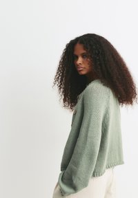 Junge Frau mit langen lockigen Haaren, die eine hellgrüne Strickjacke mit Holzknöpfen und cremefarbene Hosen vor einem weißen Hintergrund trägt.