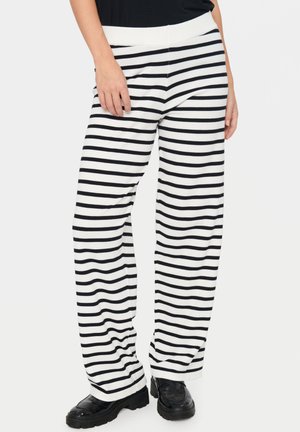 Frau trägt schwarz-weiß gestreifte, weite Hosen mit schwarzen klobigen Loafers und einem schwarzen Oberteil.