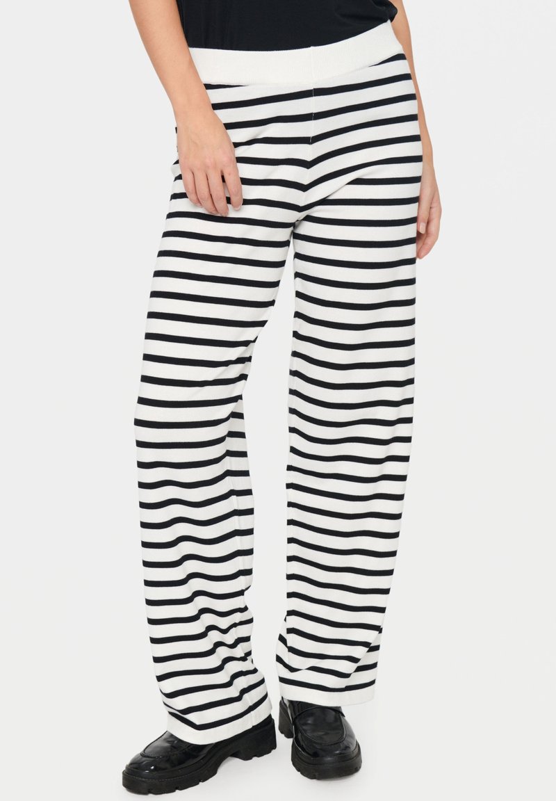 Femme portant un pantalon large à rayures horizontales noir et blanc avec des mocassins noirs épais et un haut noir.