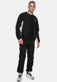 Zwarte bomberjack met witte en rode accenten, gecombineerd met zwarte broek en witte sneakers met zwarte accenten. Soepele, lichte stof.