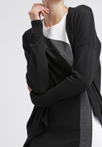 Czarny i szary kardigan z miękkiego materiału, o luźnym kroju, z długimi rękawami i otwartym przodem, noszony na białej sukience.