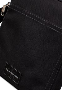 Črna platnena torba s kontrastnim belim šivom, z sprednjim žepom in pravokotno oznako z napisom "Calvin Klein Jeans" v belem besedilu.