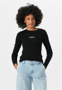 Svart langermet skjorte med ribbet tekstur og et sentralt hvitt logo på brystet, kombinert med lyseblå høytlivsjeans.