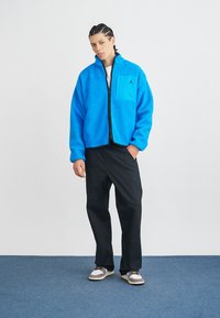 Niebieska polarowa kurtka na zamek z kieszenią; czarne spodnie; szare sneakersy z akcentami. Tło to jednolity biały kolor, z niebieską podłogą.