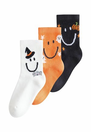 Drei Halloween-Themen Crew-Socken in Weiß, Orange und Schwarz, jeweils mit einem lächelnden Gesicht und festlichen Symbolen wie einem Hexenhut, Gespenstern und einem Kürbis.