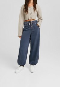 Uppskuren beige tröja med ribbat mönster och snörning framtill, tillsammans med löst sittande marinblå byxor och vita sneakers.