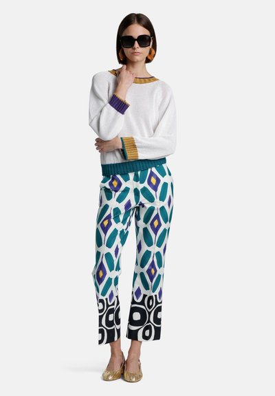 White knitted sweater with multicolored ribbed cuffs paired with patterned trousers in green, blue, and black. Gold shoes complete the look.