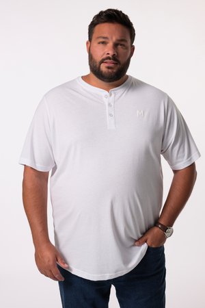 Bearded man wearing a white short-sleeve henley shirt and dark jeans, standing with one hand in pocket against a plain background.