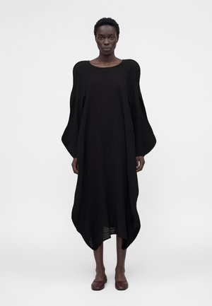 Femme portant une robe noire longue et ample avec des manches bouffantes et un ourlet arrondi, debout devant un fond clair uni, portant des chaussures rouge foncé.