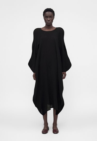 Woman wearing a loose, long black dress with balloon sleeves and rounded hem, standing against a plain light background, wearing dark red shoes.