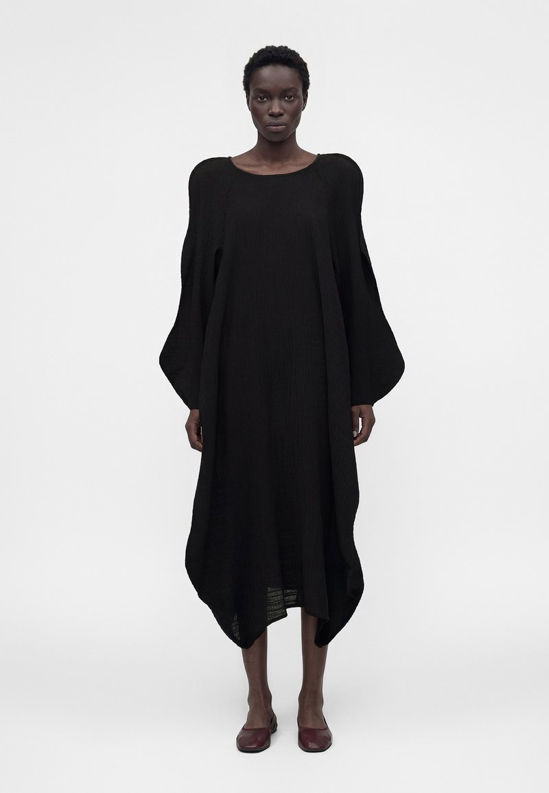 Γυναίκα φοράει ένα φαρδύ, μακρύ μαύρο φόρεμα με μπαλόν μανίκια και στρογγυλεμένη ραφή, στέκεται μπροστά σε ένα απλό φωτεινό φόντο, φοράει σκούρα κόκκινα παπούτσια.