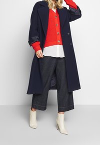 Abrigo largo navy sobre un cárdigan texturizado rojo, camisa blanca y pantalones de mezclilla oscuros de pierna ancha, completado con botas de tobillo crema.