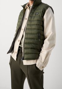 Man som bär en beige skjorta med knappar under en olivgrön quiltad väst med dragkedjefickor, kombinerad med matchande olivgröna byxor med dragsko.