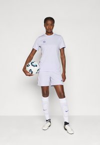 Világoslila sportmez és rövidnadrág, kontrasztos narancs részletekkel, fehér zoknikkal és stoplis cipőkkel. Egy mintás focilabdát tart.