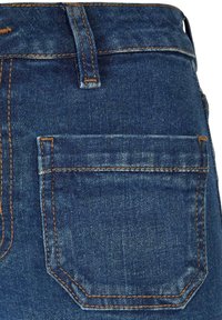 Tumedat sinised denimmid, millel on tagakott kontrastsete oranžide õmblustega ning veidi kulunud tekstuur.