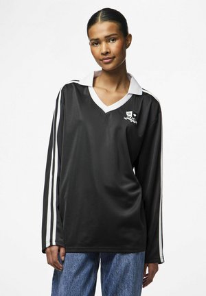 Camisa polo negra de manga larga con cuello blanco y franjas blancas en las mangas. Presenta un emblema gráfico en el pecho. Textura de tela suave.