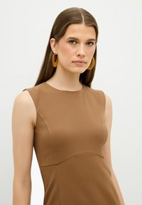 Brun ærmeløs kjole med en tætsiddende silhuet og rund halsudskæring. Modellen bærer også store cirkulære gule øreringe. Glat tekstur.