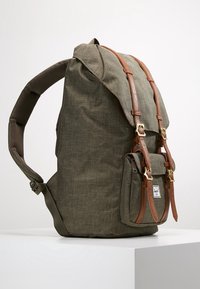 Mochila verde oliva con correas y hebillas de cuero marrón, bolsillo frontal y correas de hombro acolchadas, apoyada sobre una superficie blanca.