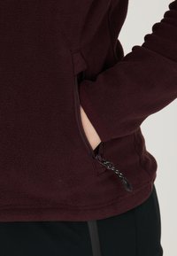 Bordeaux fleece top med en lynlåslomme på siden, med en blød tekstur og en tætsiddende design, vist sammen med mørke bukser.
