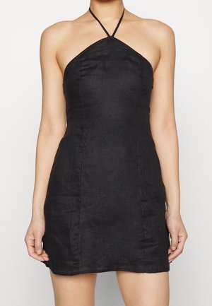 Donna che indossa un mini abito nero aderente con collo halter e dettagli cuciti, in piedi davanti a uno sfondo bianco semplice.