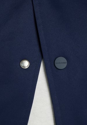 Marineblauer Stoff mit zwei Druckknöpfen; einer silberfarben, einer dunkelblau mit dem Schriftzug "new balance", der teilweise weißen strukturierten Stoff darunter zeigt.