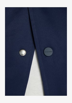 Marineblauer Stoff mit zwei Druckknöpfen; einer silberfarben, einer dunkelblau mit dem Schriftzug "new balance", der teilweise weißen strukturierten Stoff darunter zeigt.