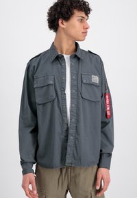 Gray shirt with button closure, two chest pockets, and a side zip pocket. Features a red tag with text. Made from lightweight fabric.