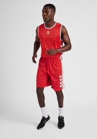 Camiseta deportiva roja sin mangas y pantalones cortos, con acentos blancos y un logo. Zapatillas de correr negras y calcetines blancos completan el conjunto deportivo.