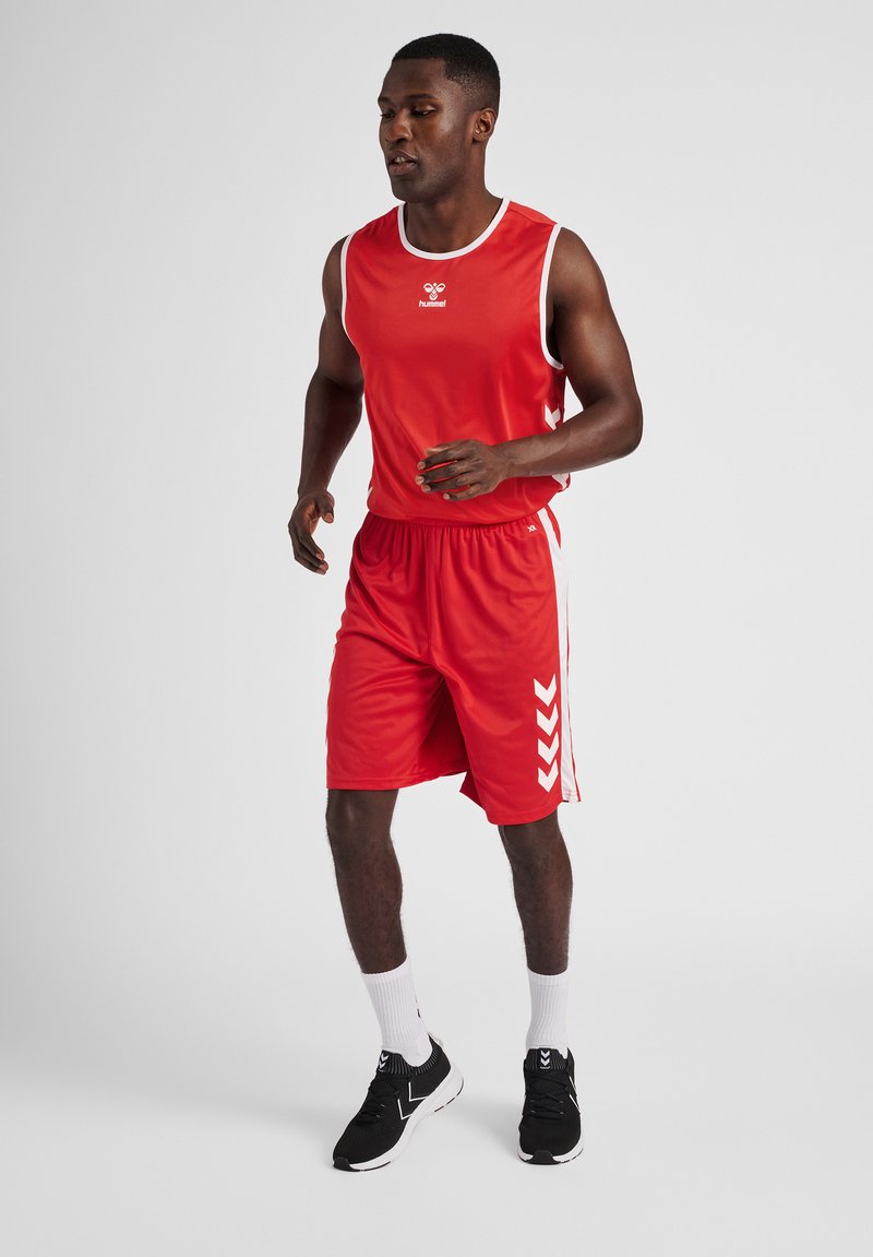 Camiseta deportiva roja sin mangas y pantalones cortos, con acentos blancos y un logo. Zapatillas de correr negras y calcetines blancos completan el conjunto deportivo.